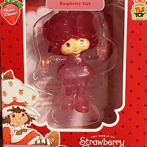 May include: A pink plastic figurine of Raspberry Tart from the Strawberry Shortcake toy line. The figurine is holding a pink bottle and has a pink dress with glitter. The packaging is red with white polka dots and features the Strawberry Shortcake logo.
