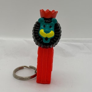 May include: A red plastic Pez candy dispenser keychain featuring a lion character head. The lion has a teal face, a black mane, a yellow muzzle, and a red crown on top. A silver metal split ring is attached to the side of the red rectangular body.