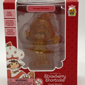 May include: A small plastic figurine of Strawberry Shortcake in a sparkly orange dress and hat. The figurine is part of a limited edition collection and is labeled "Orange Blossom".