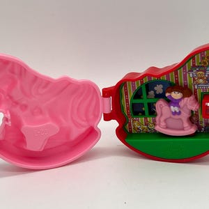 Op de afbeelding: Een vintage, roze en rode speelset in de vorm van een schommelpaard, open om het interieur te onthullen. Binnenin zit een pop op een roze schommelpaard. Het interieur heeft een raam en behangontwerp.