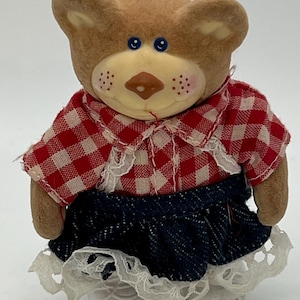 Op de afbeelding: Een bruin teddybeerbeeldje met een rood-wit geruit shirt en een spijkerbroekrok met witte kanten rand. De beer heeft blauwe ogen, een bruine snuit en kleine roze wangen. De beer draagt witte schoenen.