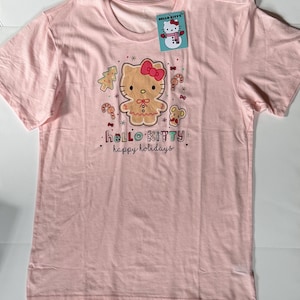 May include: A light pink t-shirt featuring a Hello Kitty gingerbread design with a red bow, surrounded by candy canes, a Christmas tree, and a mouse. The text "hello kitty happy holidays" is printed below the graphic. A Hello Kitty tag is attached.