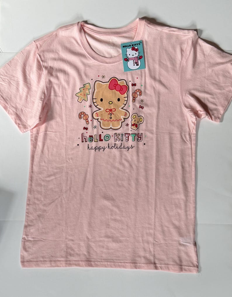 May include: A light pink t-shirt featuring a Hello Kitty gingerbread design with a red bow, surrounded by candy canes, a Christmas tree, and a mouse. The text "hello kitty happy holidays" is printed below the graphic. A Hello Kitty tag is attached.