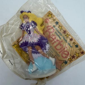 May include: A Barbie figurine from a McDonald's Happy Meal, still sealed in its original plastic packaging. The doll has long blonde hair, a purple and white dress, and is standing on a light blue base. The packaging features the words "McDonald's Happy Meal Starring Barbie."