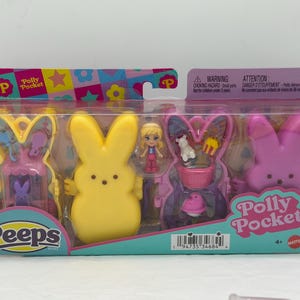 Op de afbeelding: Een Polly Pocket Peeps speelset in een doorzichtige plastic verpakking. De set bevat gele, roze en paarse konijnvormige figuren, een kleine pop en andere accessoires. De verpakking is voorzien van de Polly Pocket en Peeps logo's.