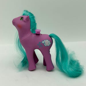 May include: A purple plastic pony toy with a bright teal mane and tail. The pony features a green eye, a red cheek blush, and a white cloud symbol on its flank. A small purple shell-shaped lever is positioned on the back of the pony figure.