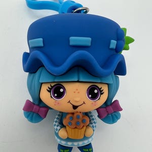 May include: Blue and purple character with a blue hat and a blue and yellow cupcake. The character has a blue dress with a white checkered pattern and a green leaf on the hat.
