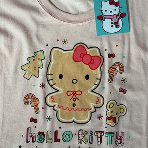 May include: A light pink Hello Kitty t-shirt with a gingerbread Hello Kitty design, Christmas tree, candy canes, and mouse graphics. The shirt has the words "hello KITTY happy holidays" printed on it. A Hello Kitty tag is attached.