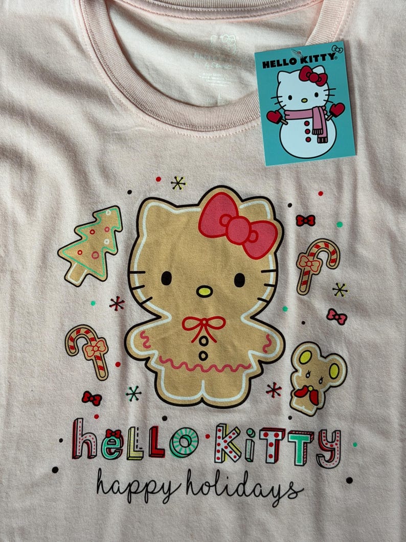 May include: A light pink Hello Kitty t-shirt with a gingerbread Hello Kitty design, Christmas tree, candy canes, and mouse graphics. The shirt has the words "hello KITTY happy holidays" printed on it. A Hello Kitty tag is attached.