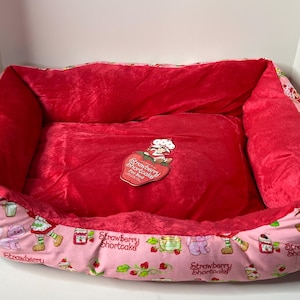 Könnte beinhalten: Ein rechteckiges Haustierbett mit rotem Plüsch-Innenraum und rosa Außenbereich. Der rosa Stoff ist mit Erdbeer-Shortcake-Figuren und Desserts gemustert. Ein Erdbeer-Shortcake-Patch befindet sich auf dem Mittelkissen.