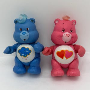 May include: Two vintage Care Bears figures. One is blue with a cloud and rain design on its belly. The other is pink with two hearts on its belly. Both have tufts of hair and heart-shaped noses.