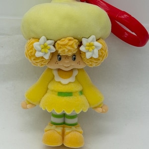 May include: A vintage, yellow plush doll with a large hat and white flower accents. The doll has a red clip and a silver ring for attachment. The doll's dress has a green stripe, and the legs have green and white stripes.
