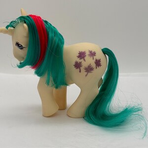 Könnte beinhalten: Ein cremefarbenes Einhorn-Spielzeug mit einer langen, fließenden türkisfarbenen Mähne, die einen einzelnen leuchtend roten Streifen aufweist. Die Figur hat ein gedrehtes Horn auf der Stirn und ein violettes Ahornblattmuster auf der Flanke.