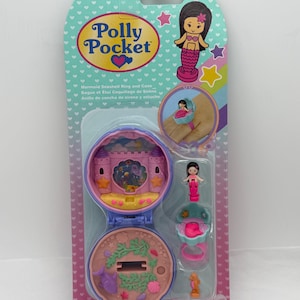 May include: A Polly Pocket Mermaid Seashell Ring and Case set. The packaging is light blue with a heart pattern and features a mermaid graphic. The set includes a ring with a miniature mermaid and a seashell-shaped case.
