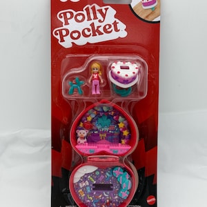 May include: A Polly Pocket playset in a heart-shaped case. The packaging is red with the Polly Pocket logo. The set includes a small doll, a cake, and a balloon animal. The case opens to reveal a miniature interior with a teddy bear.