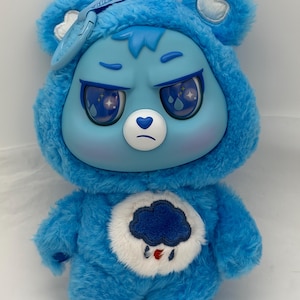 Könnte beinhalten: Ein blaues Plüschbär-Spielzeug mit grimmigem Gesichtsausdruck. Der Bär hat eine Kapuze und ein Wolkenmuster auf dem Bauch. Die Augen haben ein Regentropfen-Design. Der Bär trägt einen blauen Hut mit dem Text "Care Bear".