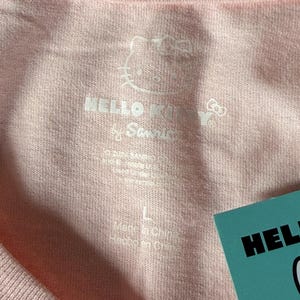 May include: Close-up of a light pink t-shirt with the Hello Kitty logo and text. The shirt has a tag that says "Hello Kitty" and the size is "L". The shirt is made in China.