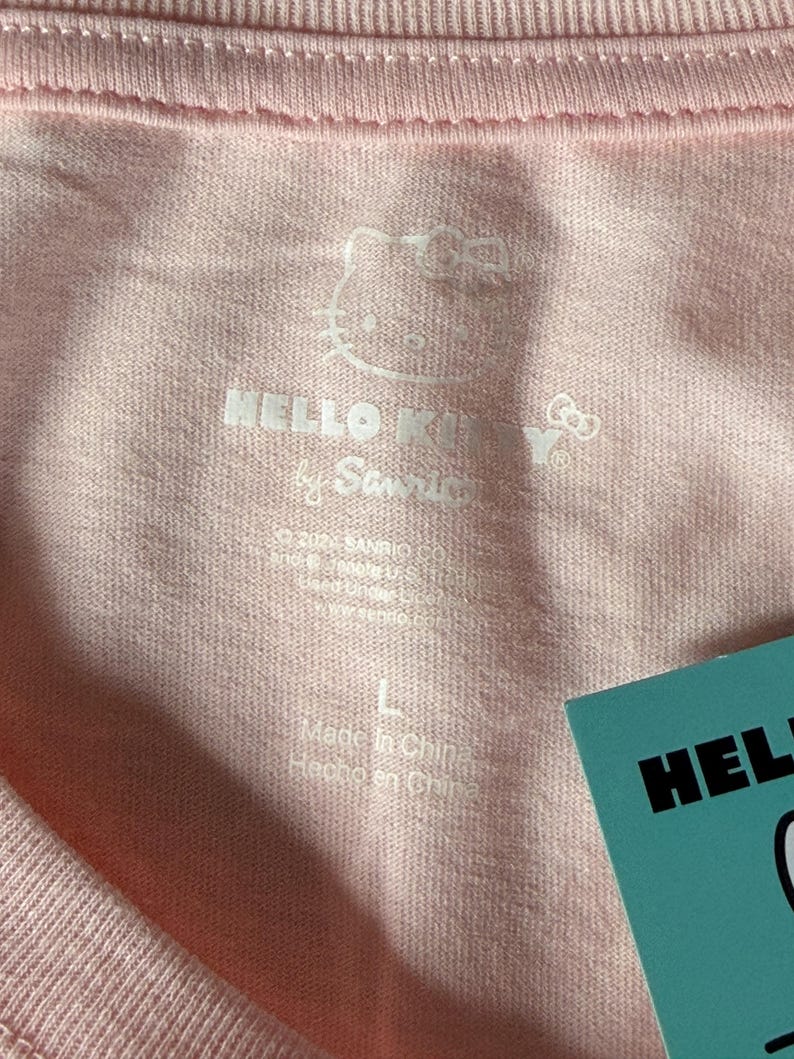 May include: Close-up of a light pink t-shirt with the Hello Kitty logo and text. The shirt has a tag that says "Hello Kitty" and the size is "L". The shirt is made in China.