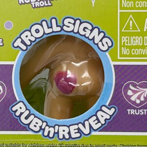 以下が含まれることがあります： 透明なプラスチックパッケージに入ったRuby Pink Trollのおもちゃのクローズアップ。パッケージには「TROLL SIGNS」と「RUB'n'REVEAL」の文字が記載されています。おもちゃの体にはピンク色のデザインがあります。背景は緑と紫です。