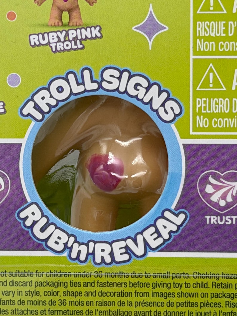 以下が含まれることがあります： 透明なプラスチックパッケージに入ったRuby Pink Trollのおもちゃのクローズアップ。パッケージには「TROLL SIGNS」と「RUB'n'REVEAL」の文字が記載されています。おもちゃの体にはピンク色のデザインがあります。背景は緑と紫です。