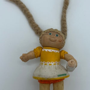 Op de afbeelding: Een kleine plastic Cabbage Patch Kid pop met blonde garenvlechtjes, die een gele jurk met witte rand en een rode broek draagt. De pop houdt een witte en groene fles vast.