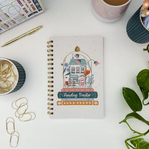 May include: A spiral-bound notebook with a whimsical illustration of a bookstore inside a glass dome. The notebook features the text "Reading Tracker" and is surrounded by other stationery items, including a gold pen and paper clips.