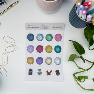 Op de afbeelding: Een vel stickers met kleurrijke maanontwerpen en kleine illustraties. De stickers bevatten verschillende kleuren zoals blauw, groen, roze en geel. De tekst op het vel luidt "12 Moon Challenge & Retelling Reading Challenge" en "www.book-sisters.com".