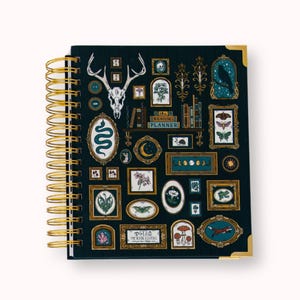 May include: A dark blue spiral-bound planner with gold wire binding and corner protectors. The cover features various framed illustrations including a deer skull, a snake, a moon, mushrooms, a fox, and text reading The Reading Planner.