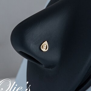 May include: A gold nose ring with a teardrop design and small clear stones. The ring is on a black display stand.
