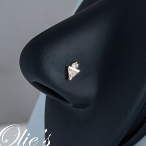 May include: A gold nose ring with a triangular design and three clear gemstones. The ring is on a black display stand.