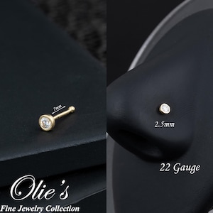 May include: A gold nose stud with a bezel-set, clear gemstone. The stud is 7mm long and the gemstone is 2.5mm. The jewelry is 22 gauge. The image includes the text "Olie's Fine Jewelry Collection."