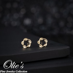 May include: A pair of gold-colored stud earrings. Each earring features a floral design with a twisted metal appearance. The earrings are displayed on a black surface with a blurred background. The text "Olie's Fine Jewelry Collection" is visible.