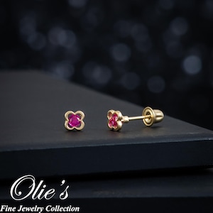 May include: A pair of gold-colored stud earrings with a four-leaf clover design. Each clover has a vibrant pink center. The earrings are displayed on a dark surface with the text "Olie's Fine Jewelry Collection".