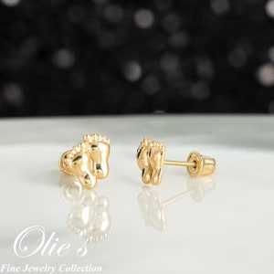 May include: A pair of gold-colored stud earrings shaped like tiny footprints. The earrings are highly polished and reflect light, with a screw-back closure. The image includes the text "Olie's Fine Jewelry Collection".