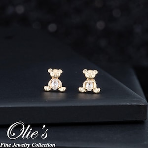 May include: A pair of gold-colored teddy bear stud earrings with a clear, oval-shaped stone in the center. The earrings are displayed on a black surface. The image also includes the text "Olie's Fine Jewelry Collection".