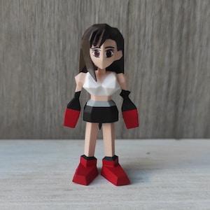 May include: A low-poly style figurine of a female character with black hair, a white top, a black skirt, and red shoes. The figurine is standing on a light wood surface.