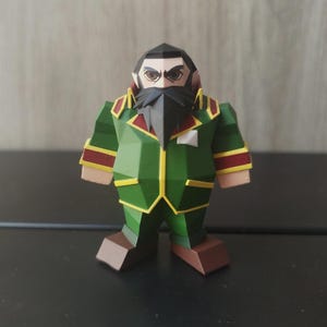 May include: A papercraft figurine of a bearded man in a green uniform with red and yellow accents. The figure has a black beard and hair, and is standing on a dark surface. The figurine is a low-poly design.