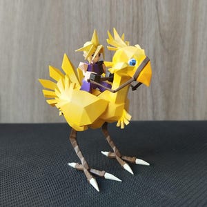 Może przedstawiać: Figurka Chocoboo wykonana w technice origami z żółtego papieru. Chocobo pochodzi z serii gier Final Fantasy. Na grzbiecie Chocoboo siedzi mała figurka Clouda Strifea.