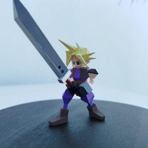 Może przedstawiać: Papierowa figurka postaci z kolczastymi blond włosami, niebieskimi oczami i fioletowo-brązowym strojem, trzymająca duży szary miecz. Postać jest ustawiona w dynamicznej pozie, gotowa do akcji.
