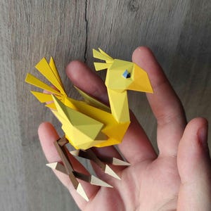 Puede incluir: Una figura de papel origami amarilla de un pájaro con patas marrones y garras blancas. El pájaro tiene un ojo azul y está en posición de pie.