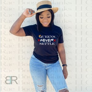 May include: A black t-shirt with the text "QUEENS XNEVERX SETTLE" in white and red. A gold crown is above the word "QUEENS". The person wearing the shirt is holding a straw hat.
