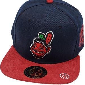 Puede incluir: Una gorra de béisbol azul marino y roja con visera plana. La gorra presenta una visera de ante roja, un botón rojo en la parte superior y un logotipo de dibujos animados de nativos americanos en la parte delantera. También tiene un emblema circular y un logotipo blanco en el lateral.