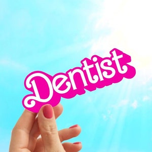 May include: A hand holds up a pink paper sign with the word "Dentist" written in white letters against a bright blue sky.