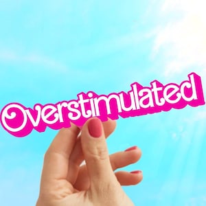 May include: A hand holds up a pink sign with the word "Overstimulated" written in white letters. The sign is held against a bright blue sky with white clouds.