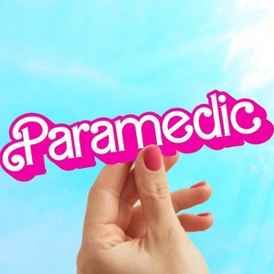 May include: A hand holds up a pink paper sign with the word "Paramedic" written in white letters against a blue sky background.