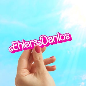 May include: A hand holding a pink sign with the text "Ehlers-Danlos" against a blue sky with white clouds.