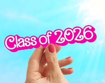 Op de afbeelding: Een roze en wit bord met de tekst "Class of 2026" wordt omhoog gehouden tegen een helderblauwe lucht met zonneschijn. Het bord wordt vastgehouden door een hand met rode nagellak.