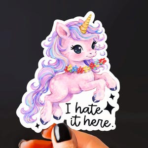 May include: A sticker featuring a pink unicorn with a yellow horn, purple and blue mane, and a floral necklace. The text "I hate it here" is below the unicorn. The sticker is held against a black background.