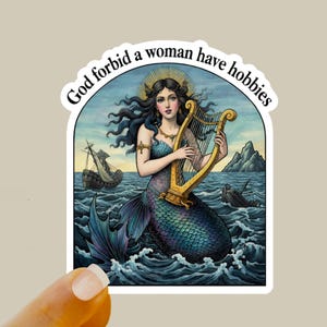 May include: A sticker featuring a mermaid playing a golden harp in the ocean. The mermaid has long dark hair and a blue tail. The sticker has the text "God forbid a woman have hobbies" in a white arch.