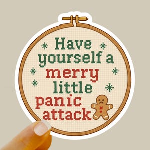 May include: A sticker with a cross-stitch design in a round embroidery hoop. The text reads "Have yourself a merry little panic attack" in green and red. A sad gingerbread man is on the right.